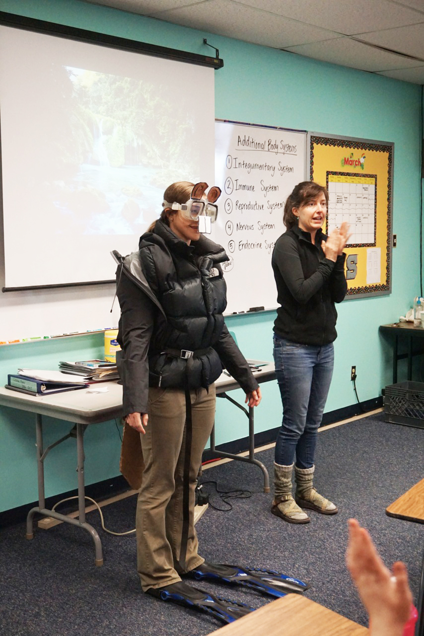 A teacher dressed as a beaver, with goggles and flippers, beside an Ecoss scientist at the front of a classroom